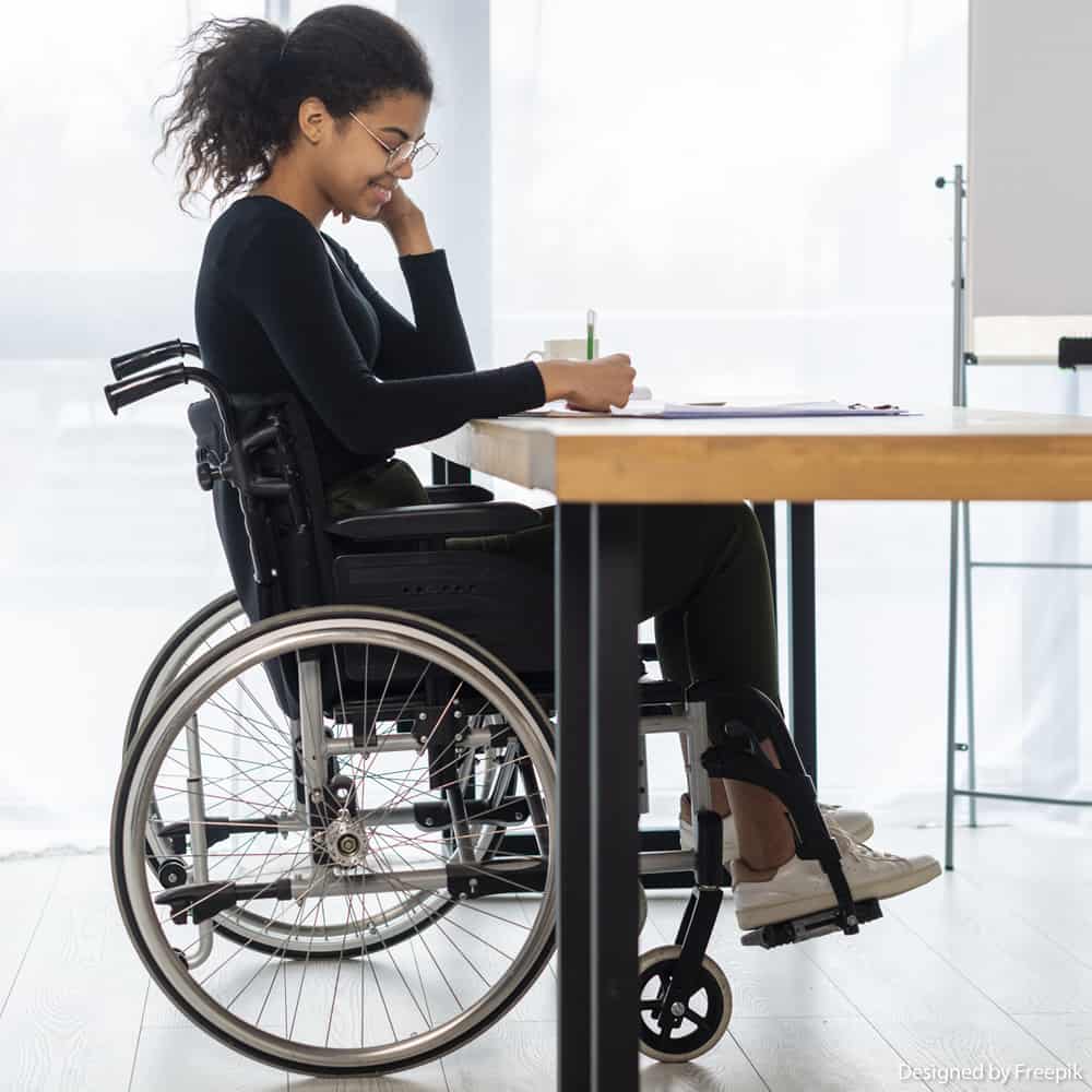 Mulher negra com deficiência física está utilizando a cadeira de rodas enquanto escreve em uma mesa de madeira, fundo branco.